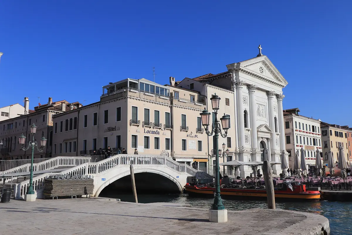 The white classical facade of the Church of the Pietà on the Venice waterfront, beside a stone footbridge over a canal